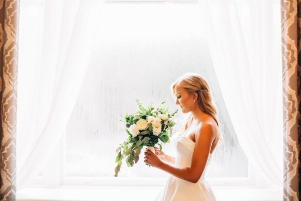 A bride stands in front of a large window and is looking down at her bouquet of flowers.